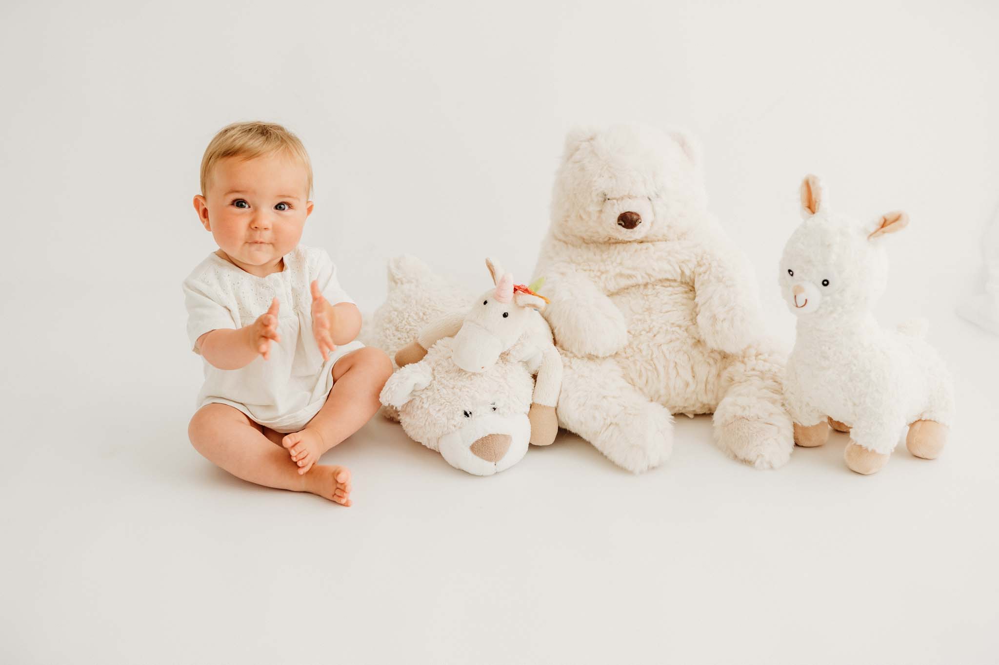 Sitzendes Baby klatscht inmitten von Stofftieren beim Baby Fotoshooting sitzendes Baby klatscht inmitten von Kuscheltieren