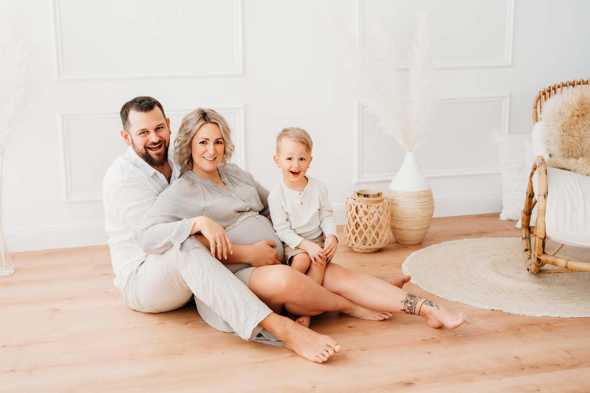Babybauchshooting Graz: Werdende Eltern sitzen mit Geschwisterkind am Boden und lachen in Kamera Familie mit werdenden Eltern und Geschwisterkind sitzen am Boden beim Babybauchshooting Graz