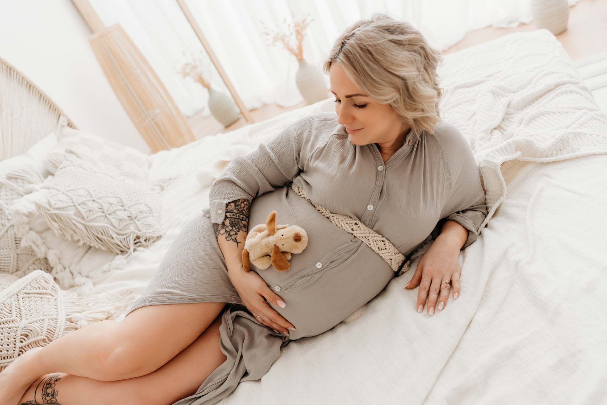 Schwangere Frau liegt beim Babybauch-Shooting Graz mit Kuscheltier am Bett im Boho Stil Schwangere Frau mit Kuscheltier am Bett